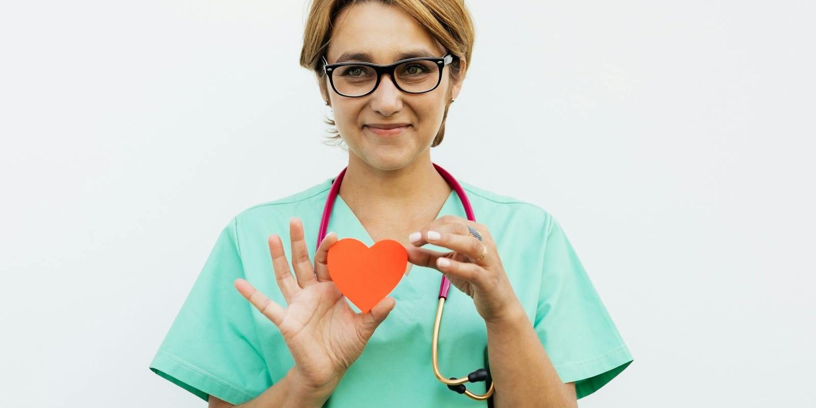 Female doctor in scrubs holding a paper heart symbol, smiling with a stethoscope around her neck.