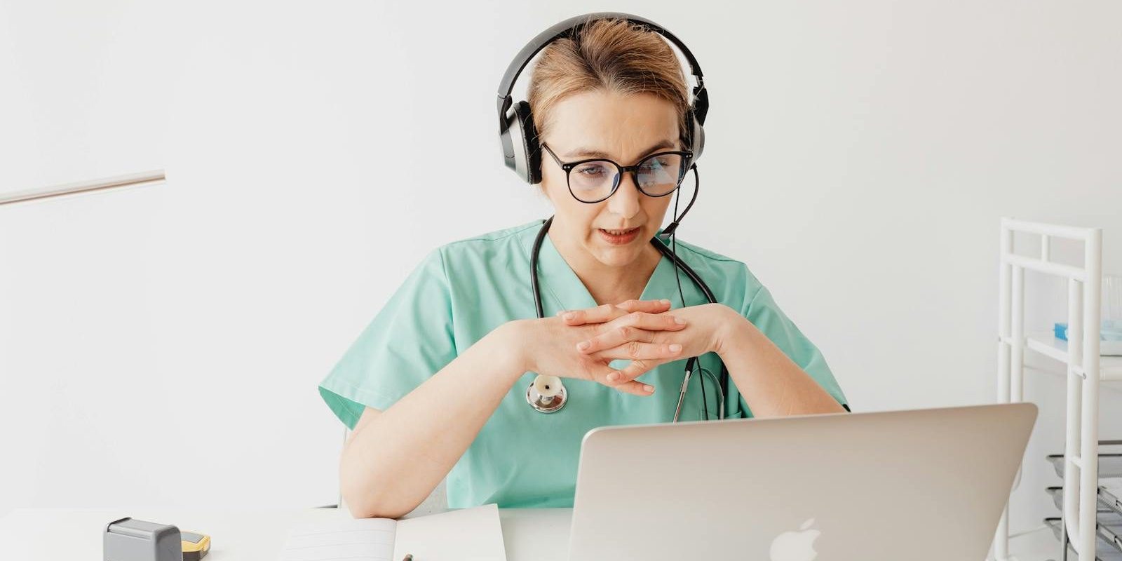 Doctor in scrubs having an online video call from her office, wearing headphones.