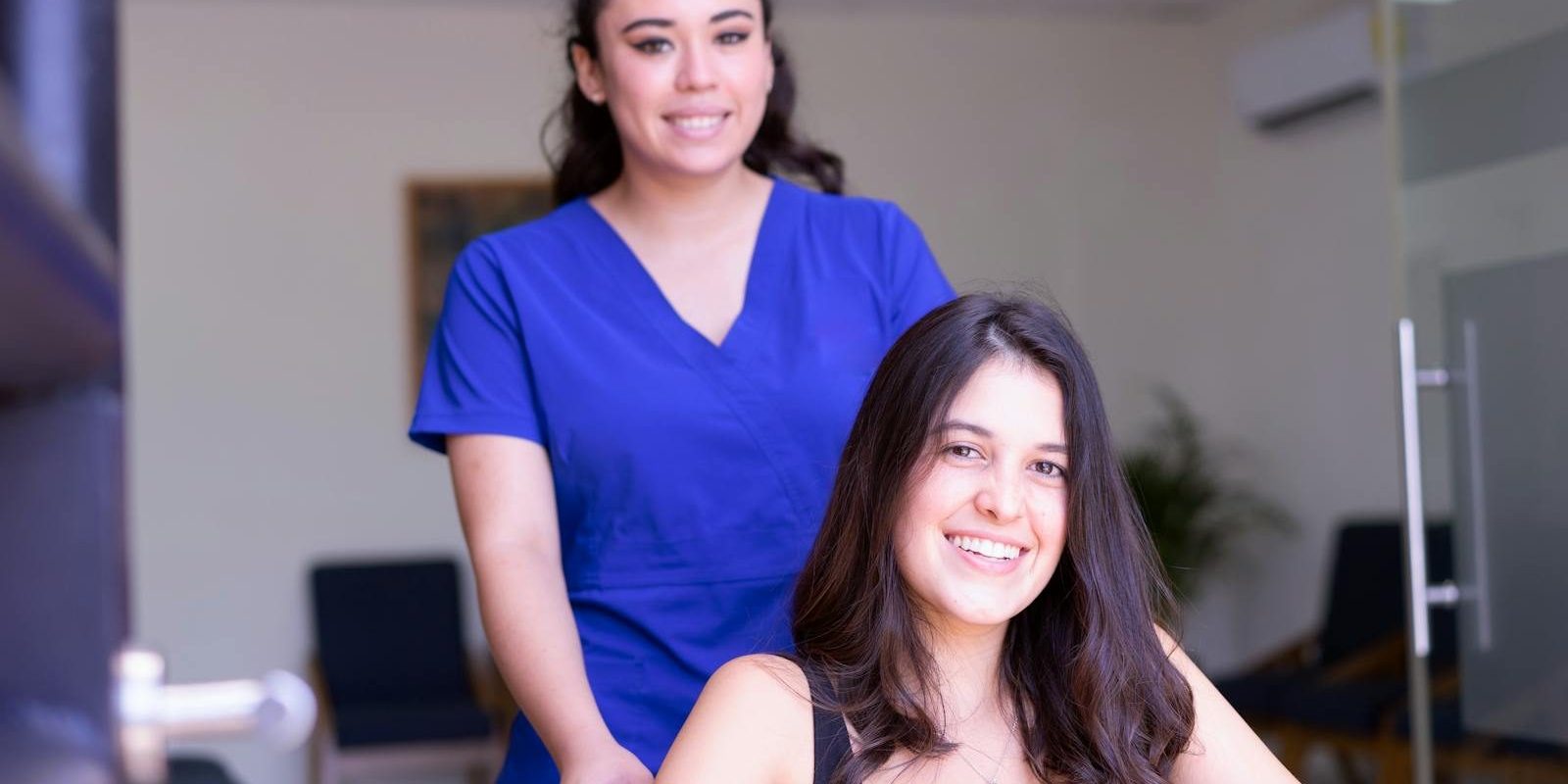 A nurse in blue scrubs assists a smiling patient in a wheelchair indoors.